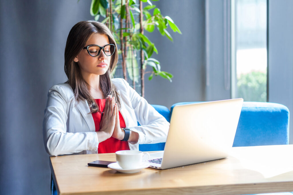 Portrait of calm beautiful stylish brunette young woman in glasses sitting and doing yoga meditation namaste pose with closed eyes and relaxed. indoor studio shot, cafe, office background.